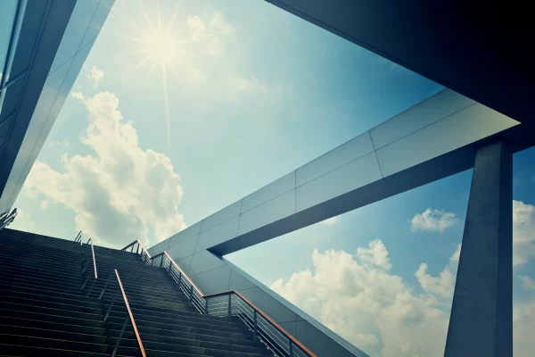 Business Coaching: Das Bild zeit eine Treppe eines Gebäudes; darüber erstreckt sich der Himmel. Die Sonne scheint und es sind wenige Wolken zu sehen. Die Treppe ist Symbol für das Erklimmen von Erfolgen.