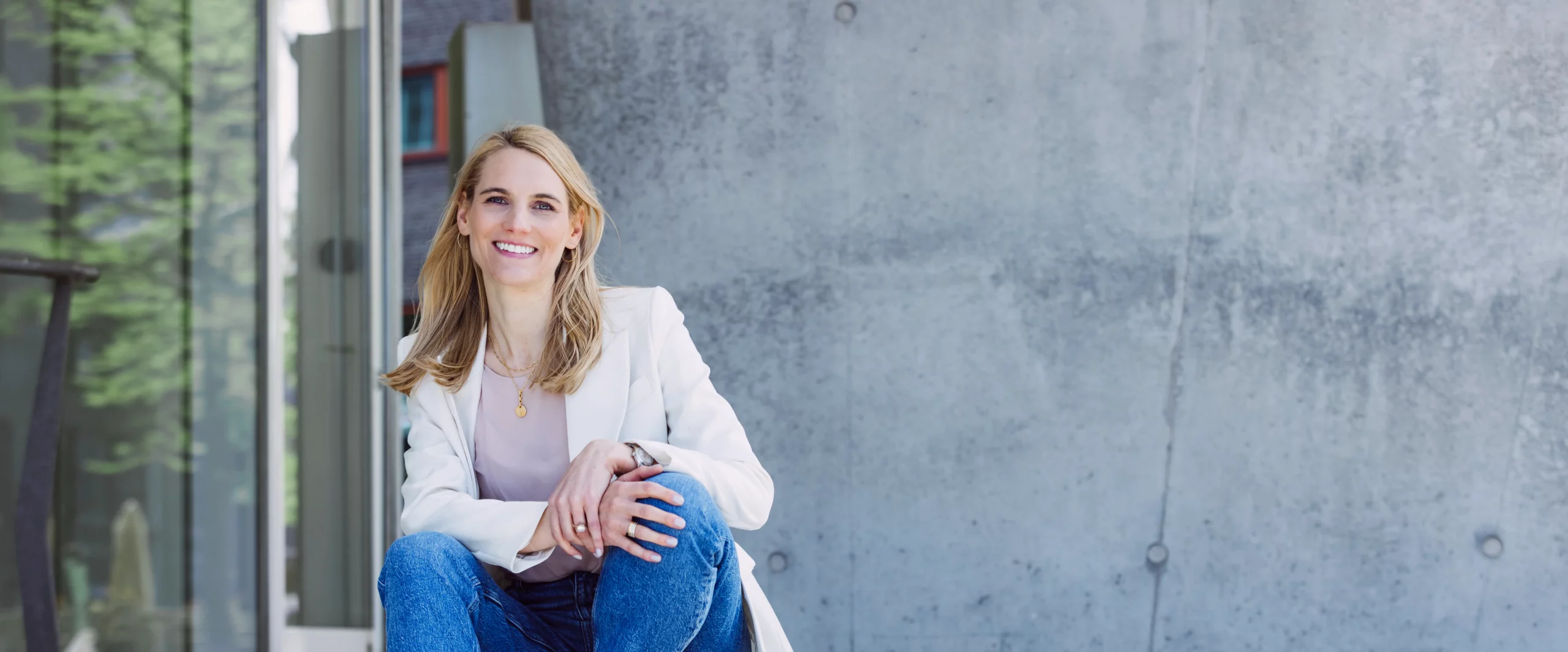 Dies ist ein Foto von Life, Business und Health Coach Dr. Kirsten Schlömer. Kirsten Schlömer sitz auf einer Treppe vor einer Betonwand und einem Fenster, in dem sich grüne Blätter spiegeln. Sie trägt einen hellen Blazer und ein Shirt in Altrosa.